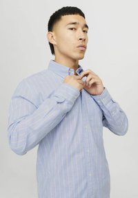 Camicia azzurra chiaro con strisce verticali bianche e gialle, maniche lunghe e colletto. Presenta una tasca e polsini abbottonati.