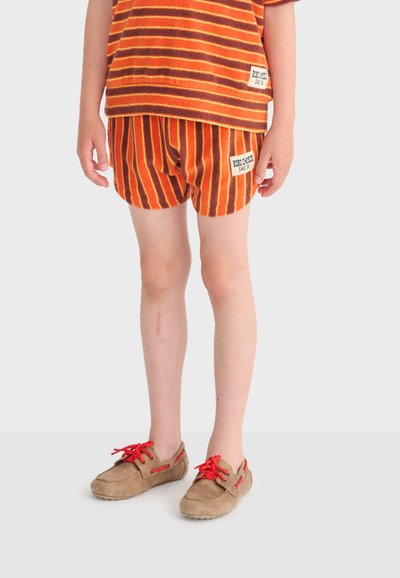 Enfant portant un short rayé orange et marron avec un haut assorti, chaussé de chaussures beige avec des lacets rouge vif, posant devant un fond uni.