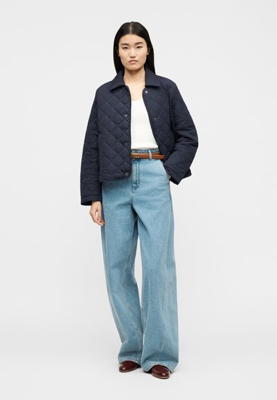 Γυναίκα που φοράει μπλε σκούρο καπιτονέ μπουφάν, λευκό τοπ, ανοιχτό μπλε παντελόνι φαρδύ με καφέ ζώνη και μπορντό παπούτσια, στέκεται μπροστά σε απλό φόντο.