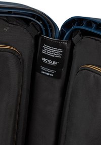 Intérieur de valise noire avec doublure en PET recyclé et coques en polypropylène, avec une fermeture éclair dorée et une étiquette détaillant la technologie des matériaux.