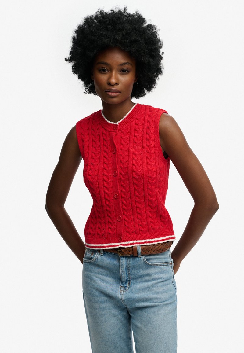 Rød strikket ærmeløs sweater med rund halsudskæring, knapper foran og hvide kanter. Modellen har lyseblå jeans og et brunt bælte på.