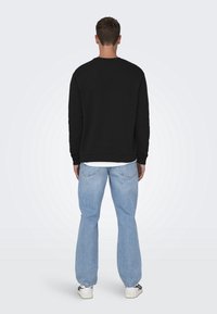 Felpa nera con collo rotondo, abbinata a jeans azzurri chiari. Il tessuto appare liscio. Sneakers con dettagli neri e bianchi sono visibili.