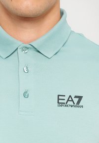 Mäta zelená polokošeľa s prehnutým golierom, tromi gombíkmi a čiernym logom EA7 Emporio Armani na ľavom hrudníku. Hladká textúra látky.