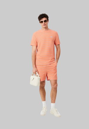 Hombre de pie con camiseta de rayas naranjas y blancas y pantalones cortos, zapatillas y calcetines blancos, gafas de sol, sosteniendo un pequeño bolso blanco.