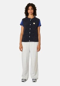 Vrouw met krullend haar, gekleed in een marineblauwe gebuttonde gebreide vest over een blauwe t-shirt, lichtgrijze broek met wijde pijpen en grijze sneakers, staand.