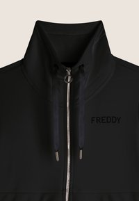 Veste noire zippée avec col montant, dotée d'une fermeture éclair argentée et de cordons noirs. "FREDDY" est imprimé en motif à pois sur la poitrine.
