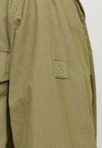 Olivgrön jacka med texturerad tyg, en lappar på ärmen och en avslappnad passform med synliga sömnadsdetaljer.