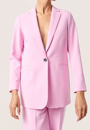 Persoon draagt een lichtroze op maat gemaakte blazer en bijpassende broek, met een enkele knoop dicht en geen hemd eronder.