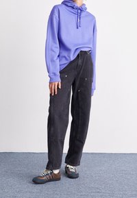 Fialová mikina, černé volné džíny s detaily ze zlaté hardware a hnědé/modré tenisky s bílými tkaničkami na modrém koberci.