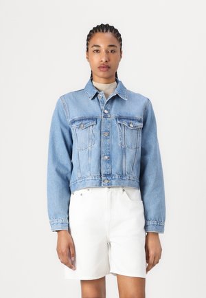 Kuttet denimjakke i lys blå med knappelukking, to brystlommer og lange ermer. Kombinert med hvite shorts.