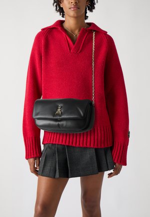 Roter gestrickter Pullover mit V-Ausschnitt und gerippten Bündchen, kombiniert mit einer schwarzen Ledertasche über die Schulter mit einem dekorativen Emblem und einer Kettenriemen.