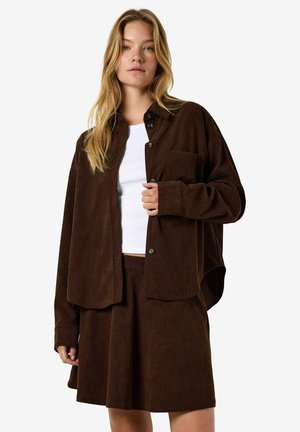 Žena s dlhými blond vlasmi, oblečená v bielom topu, hnedej oversize košeli z kordurového materiálu a zodpovedajúcej hnedej sukni na bielom pozadí.