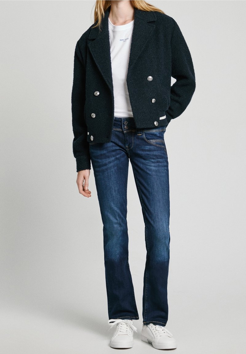 Chaqueta negra texturizada de doble botonadura con botones plateados, camiseta blanca y jeans ajustados azul oscuro, combinados con zapatillas blancas.