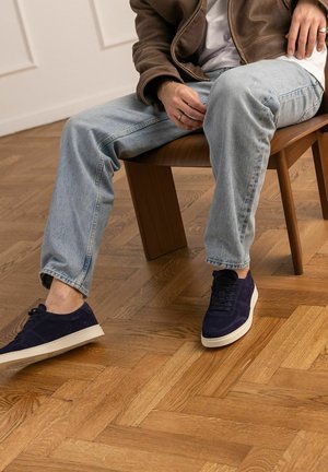 Persoon met blauwe sneakers, lichtgewassen jeans en een bruine jas zit op een houten stoel op een parketvloer.