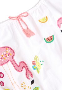 Bestickter weißer Stoff mit einem pinken Flamingo, Fruchtmotiven in Gelb und Grün sowie einer pinken Quaste als Bindung am Halsauschnitt.