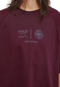 Mörkröd bomullströja med rund hals och korta raglanärmar. Har tryckta logotyper för HALO och DBU med texten "OFF PITCH."
