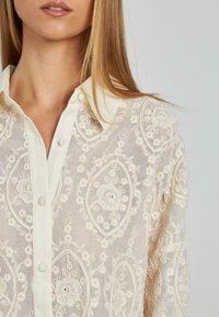 Helles beigefarbenes besticktes Blusenoberteil mit floralen Mustern, Kragen und Knopfverschluss. Weiche, transparente Textur mit kunstvollen Details.