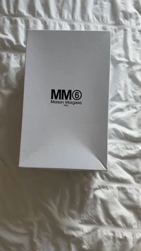 Baltas stačiakampis dėžutė su šiek tiek tekstūruota paviršiaus struktūra, viršuje juodu užrašu „MM6 Maison Margiela Paris“.