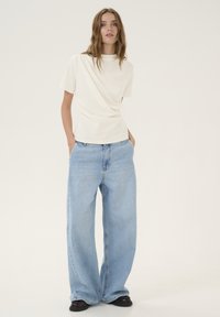 T-shirt bianco a maniche corte con scollatura alta, abbinato a jeans in denim blu chiaro a vestibilità ampia. Modella in piedi con le mani nelle tasche.