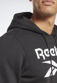 Sudadera negra de algodón con un gran logo blanco de "Reebok". Cuenta con un bolsillo frontal y capucha ajustable con cordón. Textura suave.