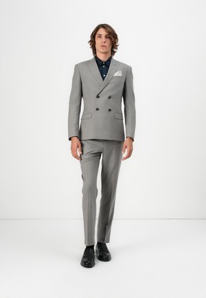 Costume gris croisé avec un motif à rayures, doté de boutons foncés, d'un mouchoir blanc dans la poche et agrémenté d'une chemise bleu marine. Chaussures noires.