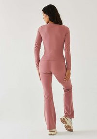 Różowy kombinezon z długim rękawem o dopasowanym kroju, gładkiej fakturze i rozszerzanych nogawkach, w połączeniu z jasnymi butami sportowymi.
