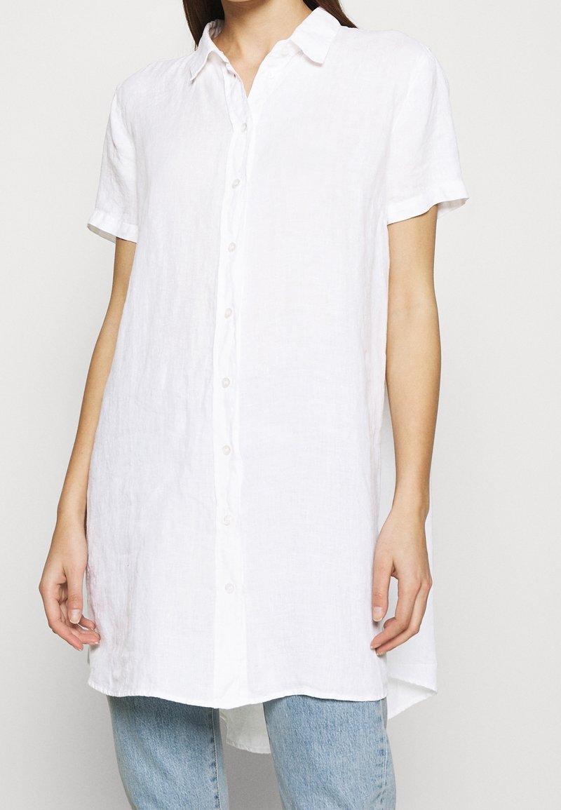 Femme portant une chemise blanche ample à manches courtes boutonnée sur un jean bleu clair, debout devant un fond uni.