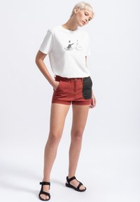 Witte t-shirt met een grafische print, gecombineerd met rode shorts met een zwart patch. Model draagt zwarte sandalen met bandjes. Eenvoudige, casual outfit.