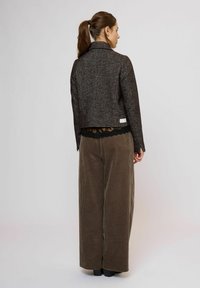 Žena s hnědým copem, oblečená v tmavém texturovaném saku, hedvábném topu s krajkovým lemem a širokých hnědých manšestrových kalhotách, stojící zády.