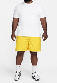 T-shirt blanche à manches courtes avec un petit logo de marque, associée à un short jaune vif. Des chaussettes blanches et des chaussures de sport noires complètent la tenue.
