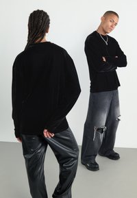 Top negru din catifea, cu maneci lungi și umeri lăsați, asortat cu pantaloni negri din piele ecologică. Modelul are părul împletit și stă în fața unui fundal deschis.