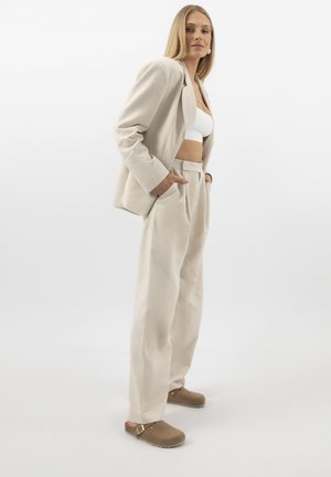 Kvinde stående med hænder i lommerne iført beige dragt, hvid cropped top og brune slip-on sko mod en ensfarvet hvid baggrund.