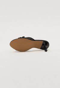 Zwarte hak sandaal met een glad plastic bovenwerk, strikaccent aan de voorkant en een getextureerde tan rubberen zool. Lage, licht gebogen hak.