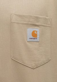 Μπεζ ύφασμα T-shirt με τσέπη στο αριστερό στήθος, στην οποία είναι ραμμένο το λογότυπο Carhartt σε γκρι και πορτοκαλί. Λείο ύφασμα, απλός σχεδιασμός.