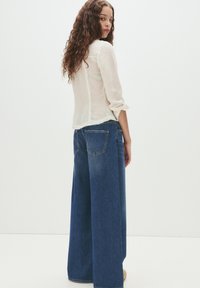 Bela teksturirana bluza z dolgimi rokavi in nabrano manšeto, v kombinaciji z širokimi modrimi denim hlačami, ki imajo zadnje žepke in udobno prileganje.