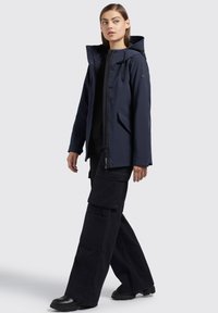 Giacca impermeabile blu navy con cappuccio, chiusura frontale con zip e due tasche laterali. Abbinata a pantaloni cargo neri larghi e stivali neri robusti.