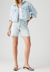 Lichtblauwe cropped spijkerjas met opgerolde mouwen, gecombineerd met bijpassende shorts met hoge taille. Zwarte sandaalhakken met bandjes maken de look compleet.