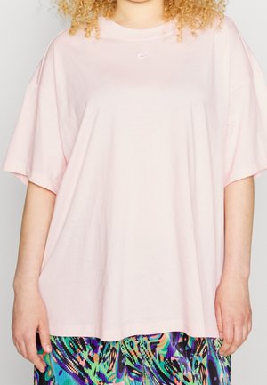 Femme portant un t-shirt ample Nike rose clair et une jupe à motifs abstraits colorés, debout devant un fond uni.