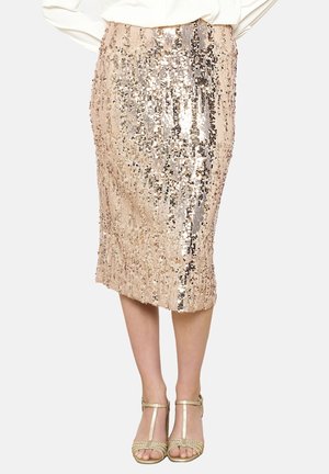 Jupe crayon longueur genou en sequins or rose portée avec des talons à brides en T dorés métalliques et un chemisier blanc rentré.