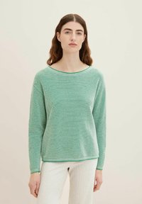 Hellgrüner, texturierter Strickpullover mit weitem Ausschnitt und langen Ärmeln, kombiniert mit ribbed crèmefarbenen Hosen, die grüne Akzentstreifen besitzen.