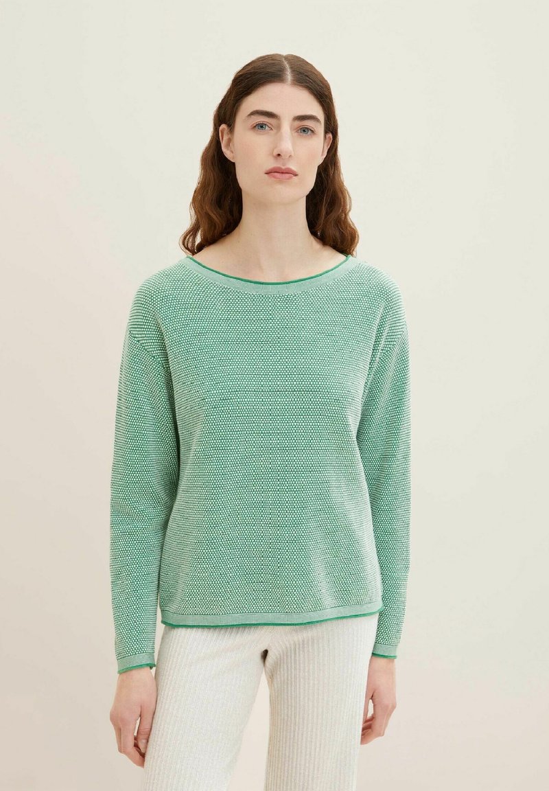 Hellgrüner, texturierter Strickpullover mit weitem Ausschnitt und langen Ärmeln, kombiniert mit ribbed crèmefarbenen Hosen, die grüne Akzentstreifen besitzen.