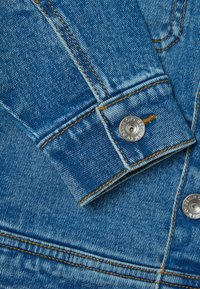 Nahaufnahme einer Jeansjacke aus leicht blauem Stoff mit verblasster Struktur, kontrastierenden orangen Nähten und einem silbernen Knopfakzent.