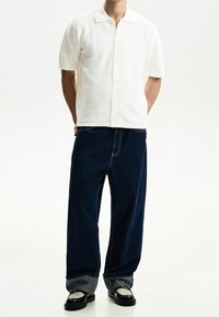 Weißes kurzärmeliges Hemd mit Knöpfen und strukturiertem Muster, kombiniert mit dunkelblauen, weiten Jeans mit kontrastierender Naht und umgeschlagenen Bündchen.