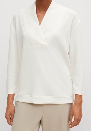 Personne portant un haut blanc à manches longues avec un large col en V et un pantalon beige, montré du cou aux hanches.