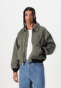Jaqueta bomber verde com gola, fecho de zip e punhos canelados pretos. Usada sobre uma camisa branca e combinada com jeans de denim azuis.