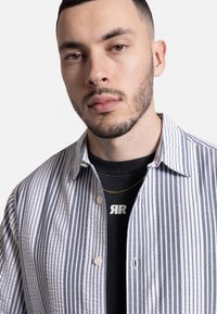 Camisa de botones con rayas verticales grises y blancas, de tela suave y con cuello puntiagudo. Llevada sobre una camiseta negra con un logo blanco.