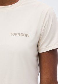 Κοντινή λήψη μιας μπεζ κοντομάνικης μπλούζας με το λογότυπο "NORRØNA" και ένα μικρό έμβλημα ζώου στο στήθος, που φοριέται από ένα άτομο με σκούρο δέρμα.