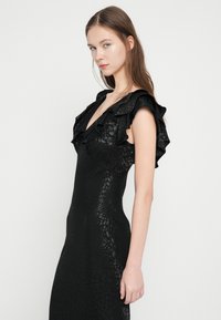 Juoda suknelė su raukšlėtu kaklo iškirpimu ir aptempta silueto forma. Tekstūruota medžiaga su subtiliu raštu. Berankovė, elegantiška ir grakšti.