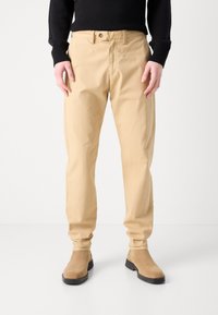 Bežové bavlnené nohavice so slim fit, s predným zapínaním na gombík, bočnými vreckami a hladkou textúrou. Nosené s hnedými semišovými čižmami.