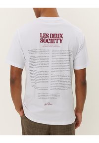 White T-shirt featuring a large graphic print on the back with text and "LES DEUX SOCIETY" in burgundy. Short sleeves, standard fit.